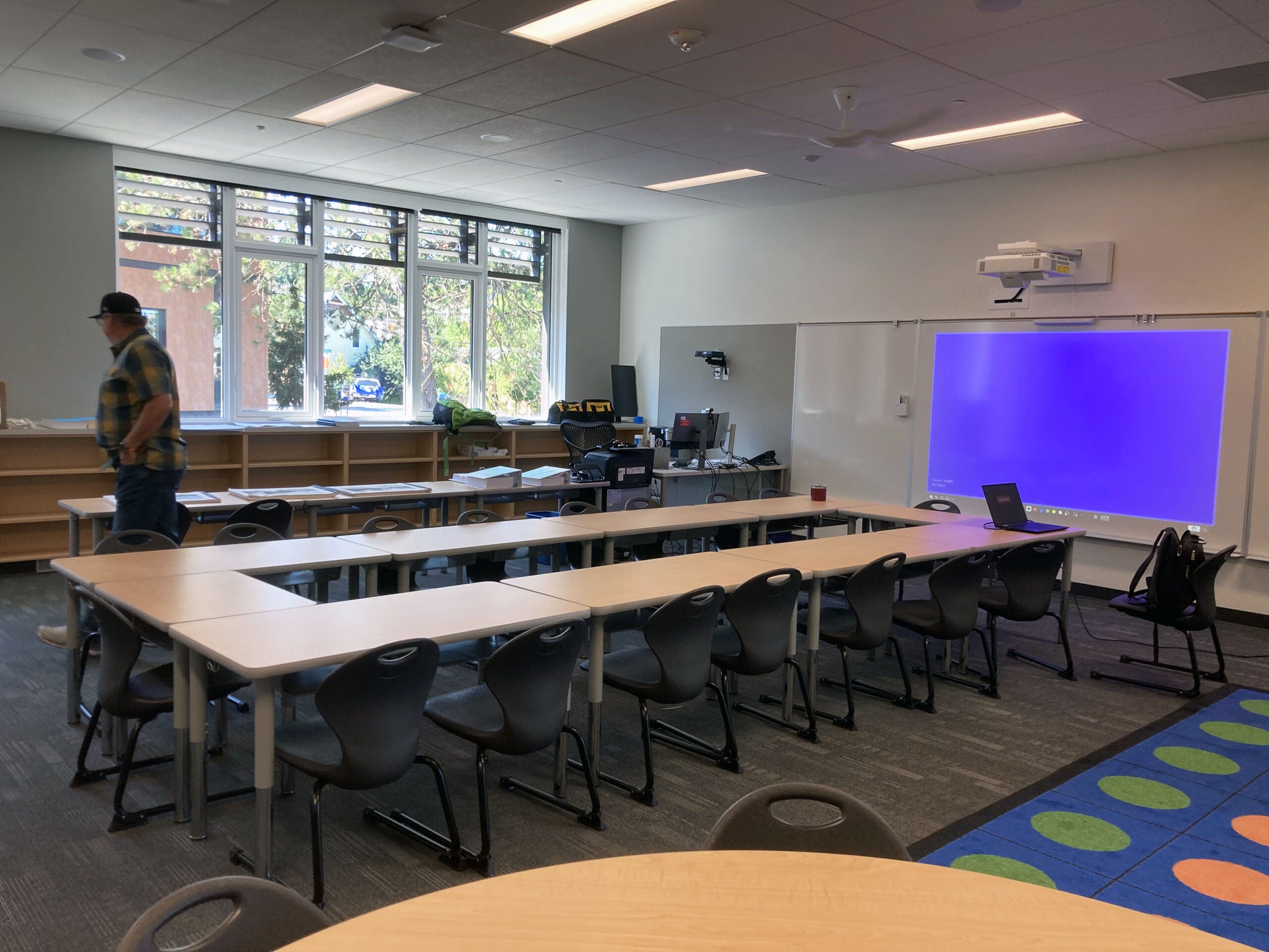 a classroom with tables set in a circle, windows on one wall with bookshelves below, a white board and projector on another wall