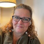 photo of Mikelle, wearing a dark green shawl or wrap, seated indoors near a beige wall with a warm-toned lamp above. The individual has curly, light brown hair and is wearing dangling earrings with decorative charms and orange accents.
