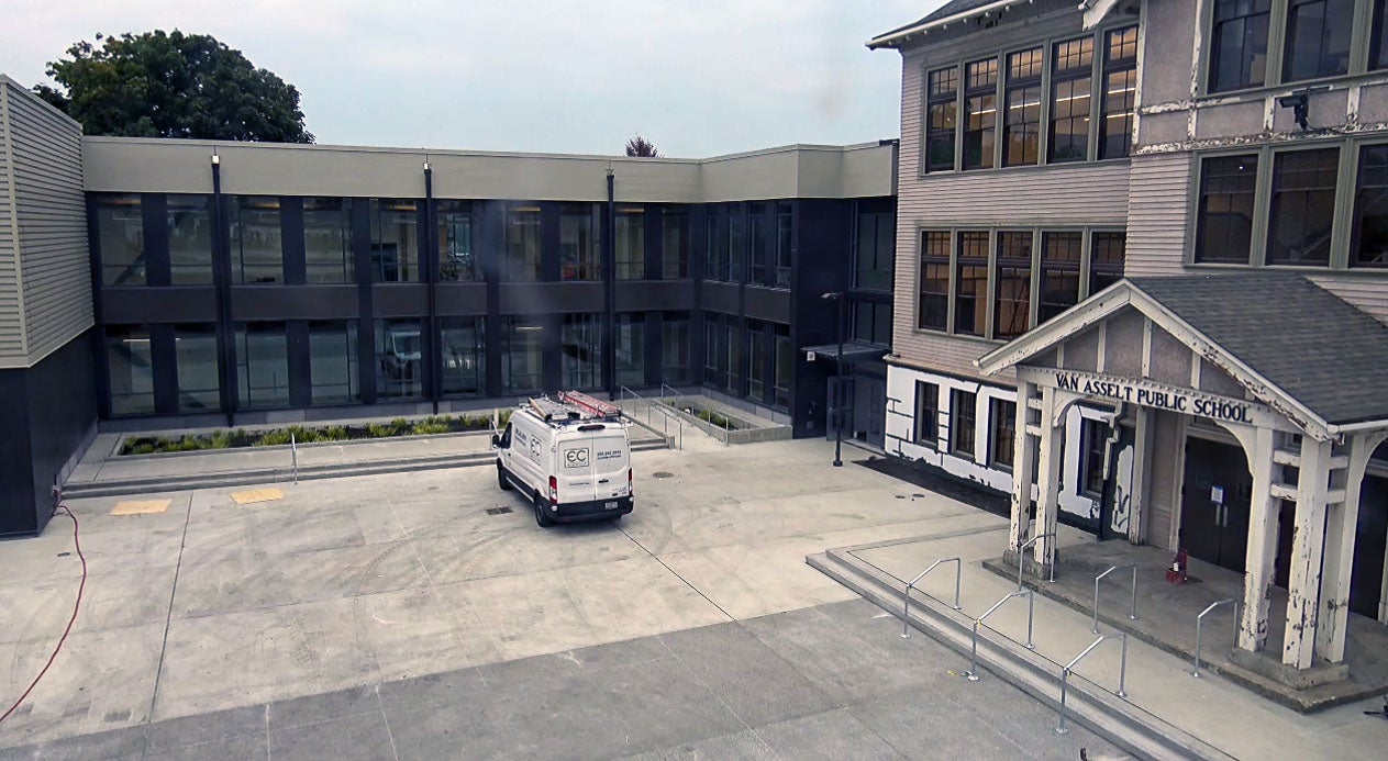 a three story historic building sits to the right with a new 2 story building directly in center with paved area in front of both