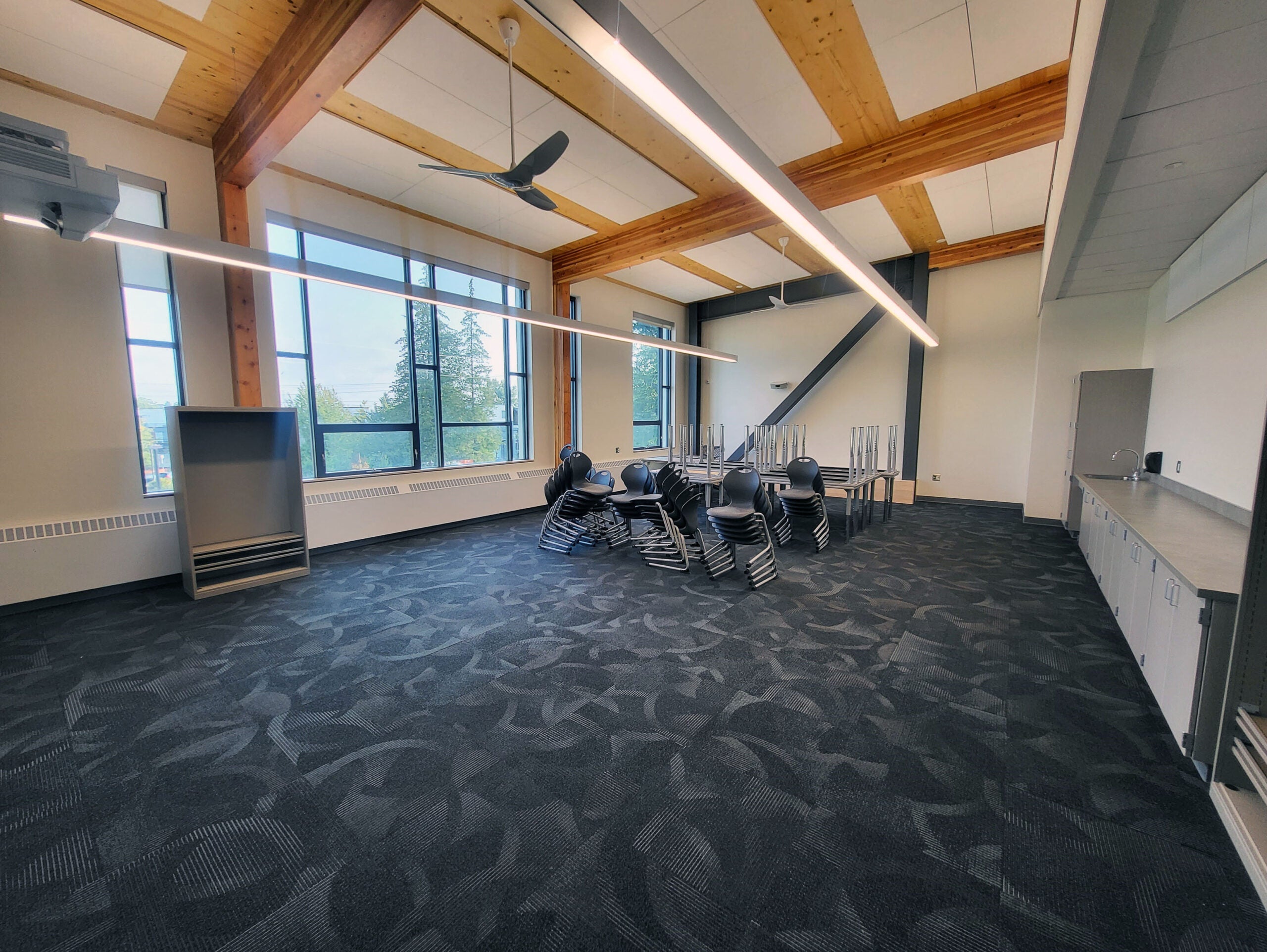 a classroom has windows on one side, wood beams in the ceiling, and a counter across from the windows - chairs and tables are stacked in the center