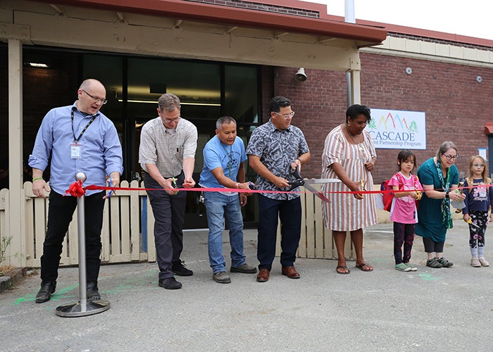 a group of people are lined up behind a red ribbon. all hold scissors with the person in the middle holding giant scissors