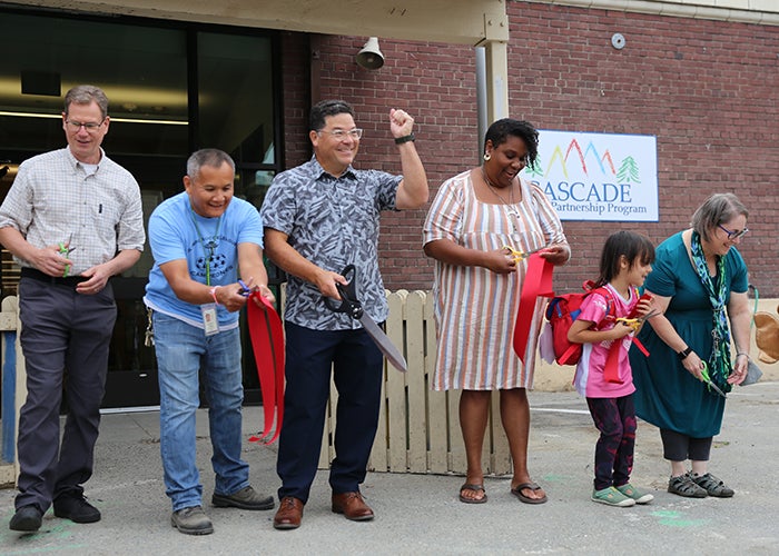 a group of people hold pieces of red ribbon. one person has huge scissors and has his hand in the air in celebration