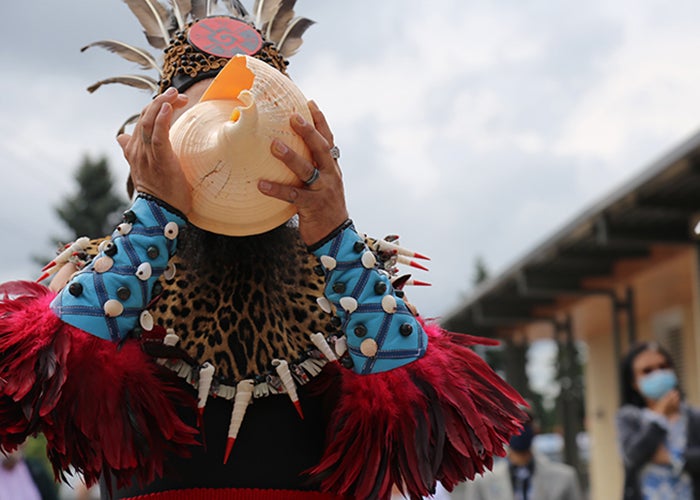a person with a feather headdress, beaded cuffs, and a necklace with teeth holds a large pink shell in front of their face