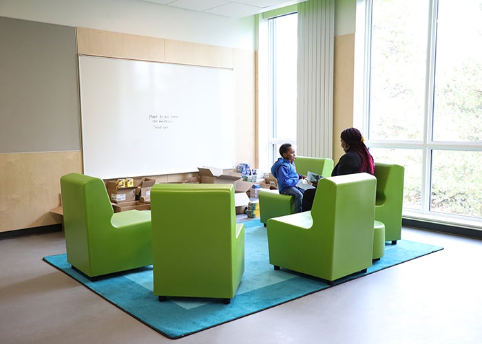 a group of lime green chairs sites on a turquoise rug in a corner with windows on both sides. an adult and a child site in the two of the seats