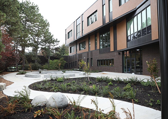 a paved walkway leads to a patio area that has seating. a three story building is behind it
