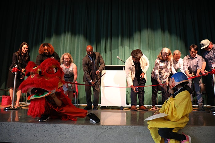 a group of people on a stage lean toward a red ribbon with scissors in their hands