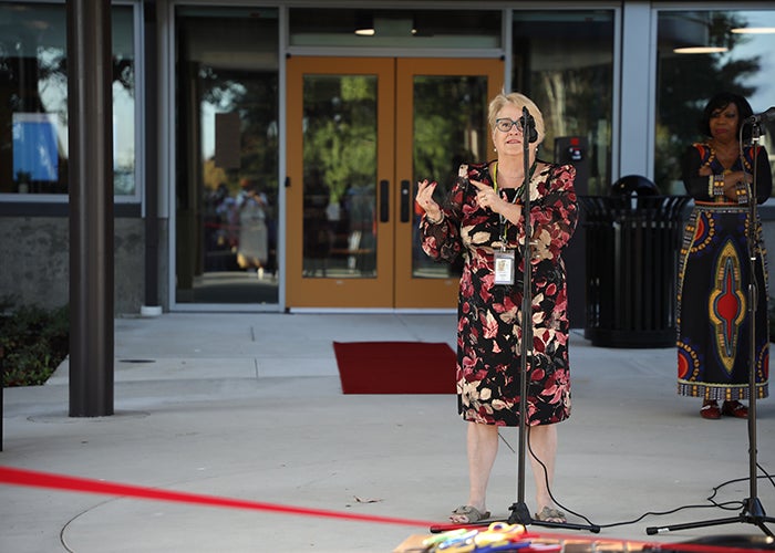 a woman in a maroon dress with pink flowers stands in front a microphone behind a red ribbon