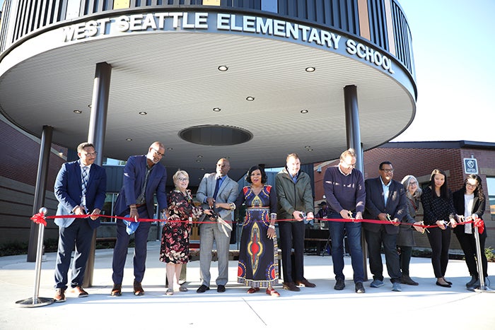 a group of people line up behind a red ribbon. all hold scissors. a circular canopy is above and behind them