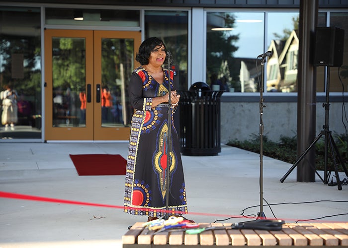 a woman in a long blue dress with red and gold figures stands in front of a microphone behind a red ribbon