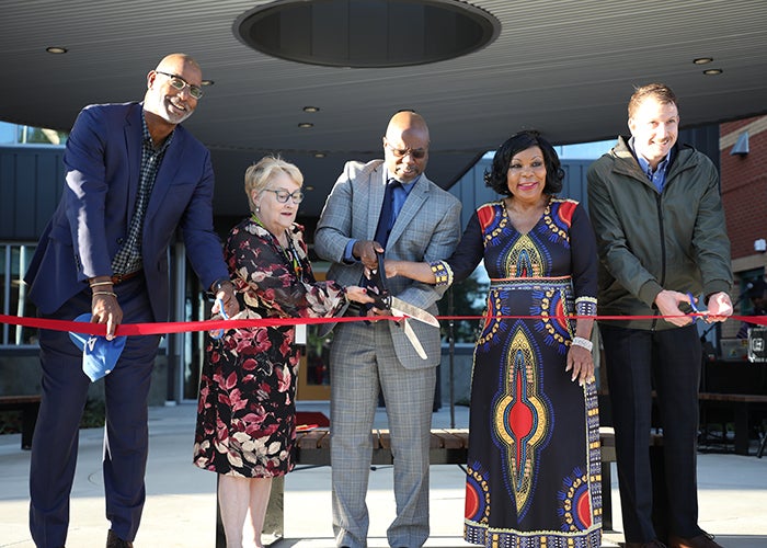 five people are behind a red ribbon, two have normal size scissors, three have their hands on a huge scissors