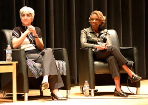 Two women sit together on a stage during a school event.