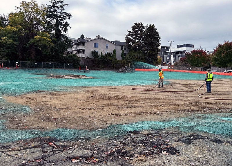 2 workers in construction vests and hats hold hoses while standing in a dirt field. part of the field has a green material on it.