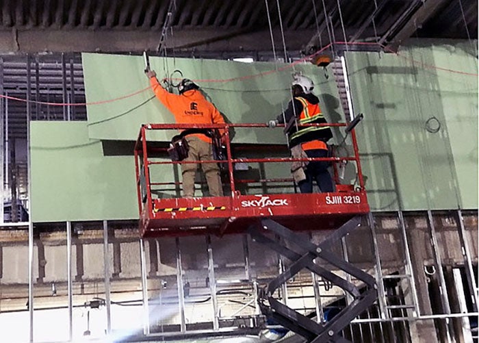 two workers in construction safety equipment are on a lift holding green sheets of material against studs