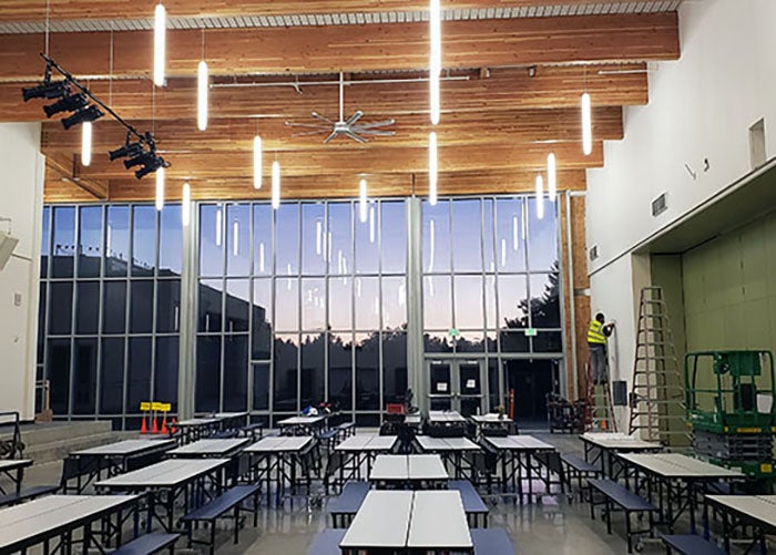 a large room with a wall of windows and exposed wood beams a stage is on the left and there are lunch tables