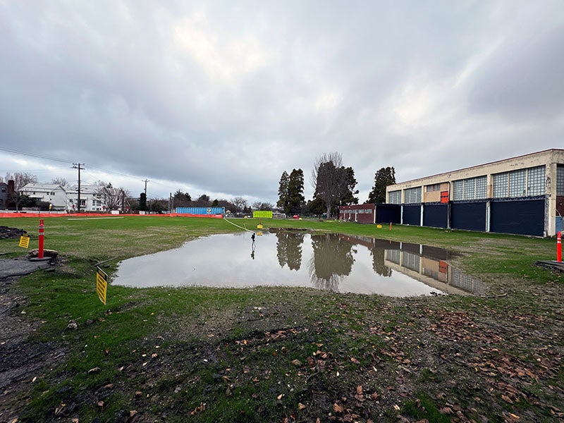 a pond of water sites amid grass near a 2-story building. there is an orange construction fence visible.