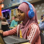 Student with headphones on smiles while working on a laptop.