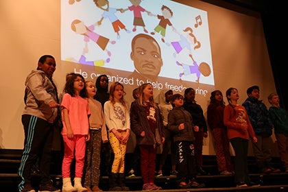 A group of John Rogers students sing in front of a projector with Dr. Martin Luther King Jr on it.