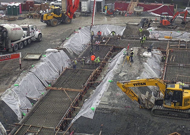 an excavator sites on dirt with a trench that has wide concrete forms with rebar and concrete pump nozzle leading into it.