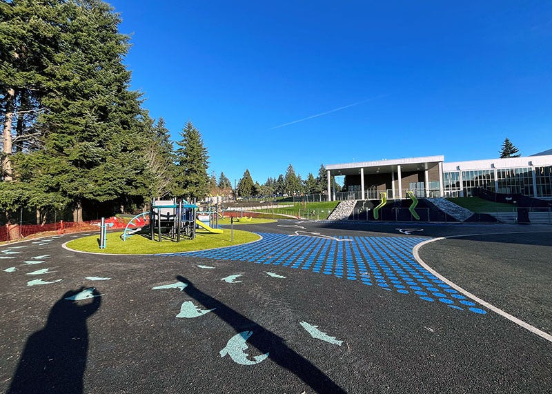 outdoor play area with a play structure, fish and filled circles painted on paving, and two slides on a slope from a building to the play area