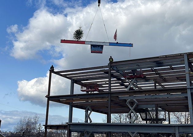 a steel beam is being slung into place at the top of a steel framed building by a crane . Two people wait on top of the existing structure