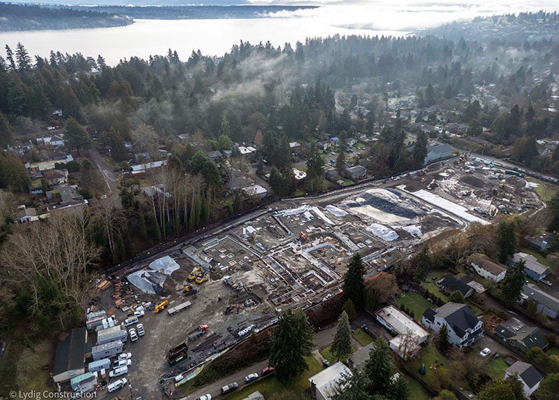 aerial view of a large construction site with equipment and trucks. there is a body of water in the background and houses in the foreground