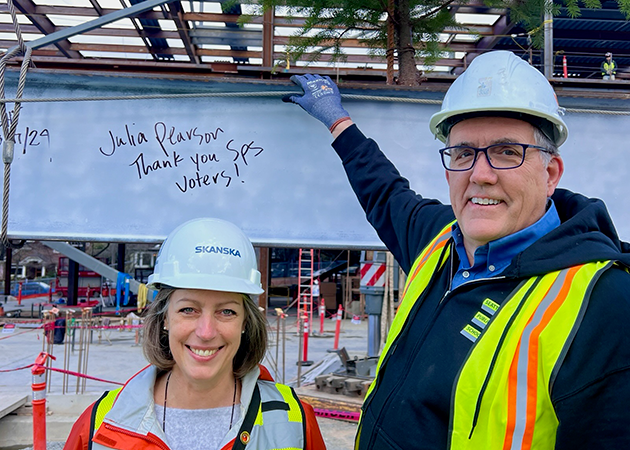 a short person and a tall person wear construction safety vests and hard hats while standing in front of part of a steel beam painted white. handwriting on the beam says "thank you SPS voters, Julia Pearson"