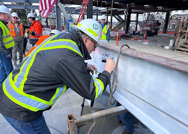 a person in a safety jacket and hard hat leans forward to write on a white painted steel beam