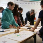 Students and elders stand around desks covered in artifacts, discuss objects.