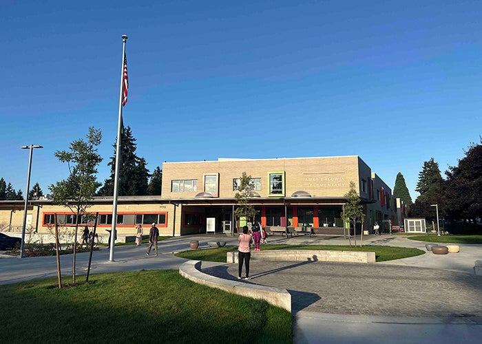 the front of a building has one part that is 2-stories and 1 part that is 1-story. it is light colored with a roofed entry. a circular paved area, an area of grass, a concrete walkway, and a flag pole are in front. there are a few people facing away from the camera