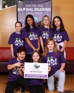 Seven students stand together in front of a sign that says Take the Global Reading Challenge 2024