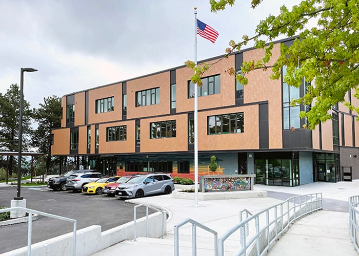 a 3 story building made of reddish bricks with the 2nd floor forming a canopy over the first floor entrance. a concrete walkway and parking lot are in front. there is an American flag on a pole.