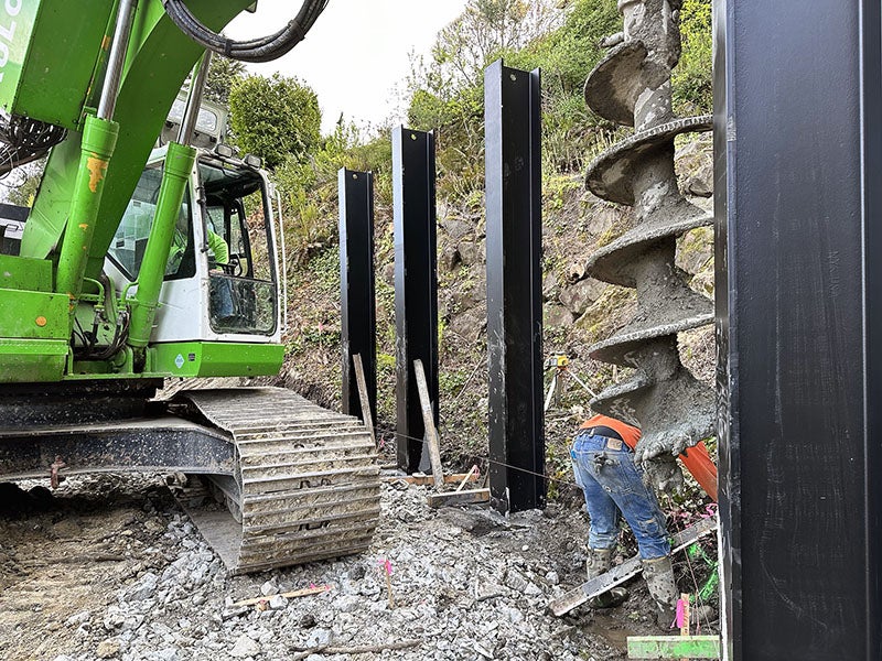 a large construction machine with tracks has a giant drill bit and is screwing it into the ground between two metal posts