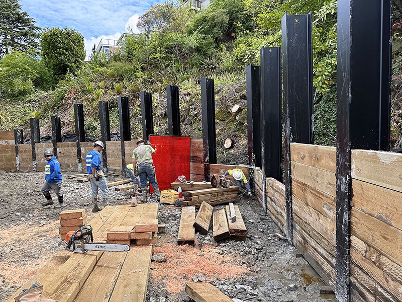 large metal posts approximately 12 feet tall are lined up along a hillside and workers are putting giant wood slabs between them