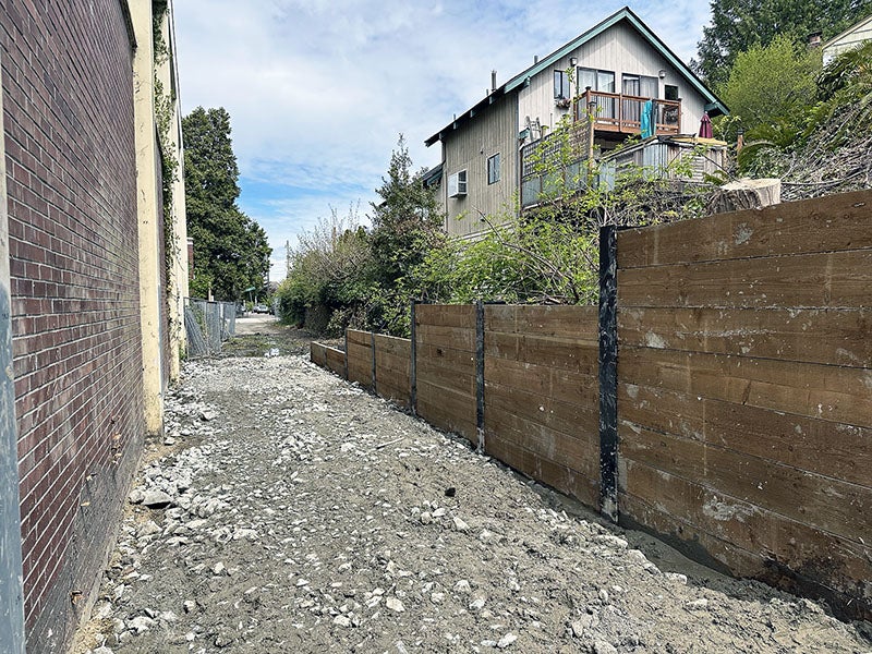 a brick wall on the left faces a partial wall made of steel posts and wood slabs on the right. a house is visible on the hill behind the right side wall