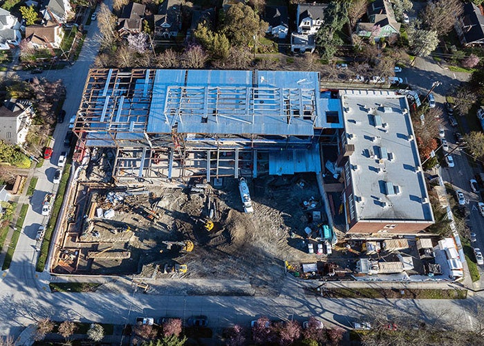 aerial view of a construction site with a brick building to the right and a new building being constructed at a right angle attached to it in the upper part of the photo. dirt and construction equipment are in the remaining space