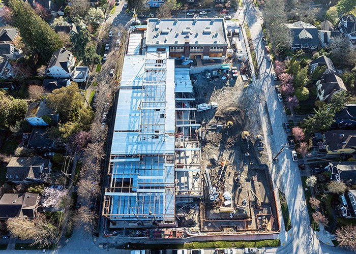 aerial view of a construction site with a brick building at the top and a new building being constructed at a right angle on the left side. a foundation is in progress in the lower right corner of the site  dirt and construction equipment are in the remaining space