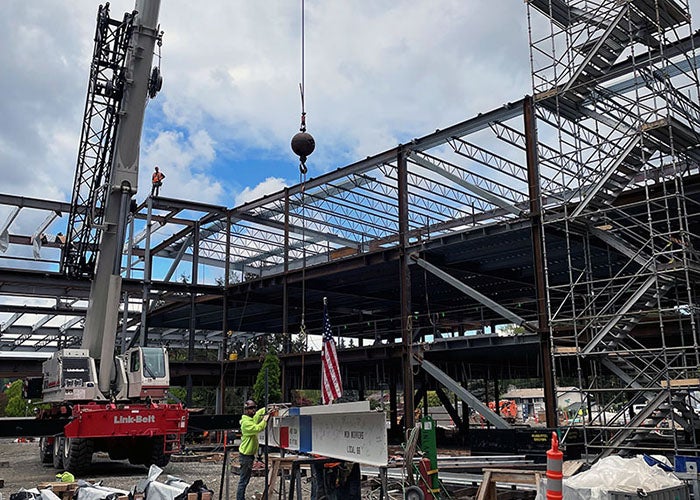 a crane line is being attached to a white steel beam with red and blue stripes. there is a US flag and an evergreen tree attached to the top of the beam