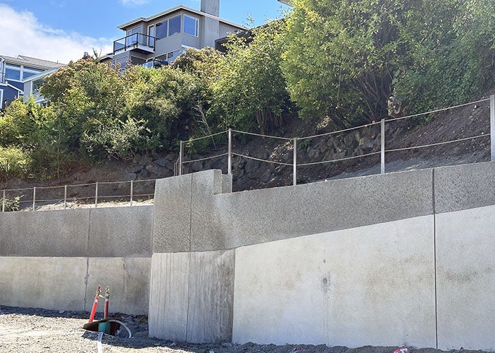a two-level concrete retaining wall has a railing on top and there is shrubbery above and houses visible above that