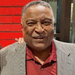 Photo of Ammon McWashington, wearing a gray checkered blazer over a maroon collared shirt, standing in front of a red brick wall with a dark window frame in the background.