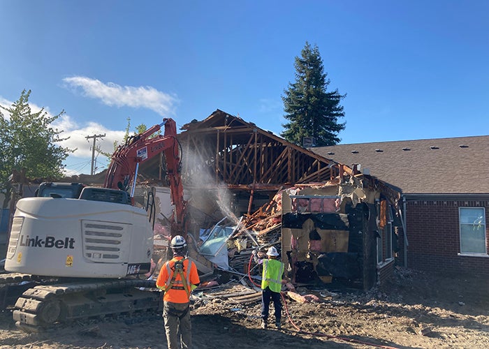 a bucket loader is demolishing a portion of a brick building while workers spray water
