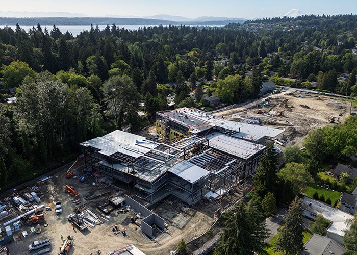 aerial view of a construction project with a large H-shaped building in progress. Behind it is a open dirt area with construction equipment. Trees are all around and a lake is visible in the far background.