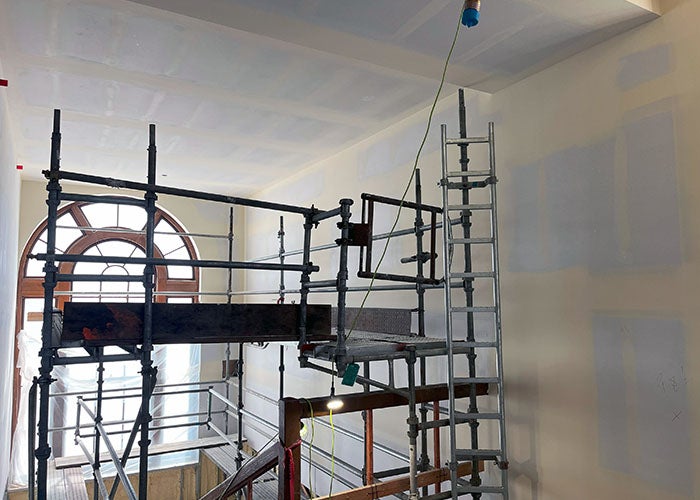 interior of a large space with high ceilings and an arched window. a scaffolding is set up and the walls have raw wallboard that is taped and mudded
