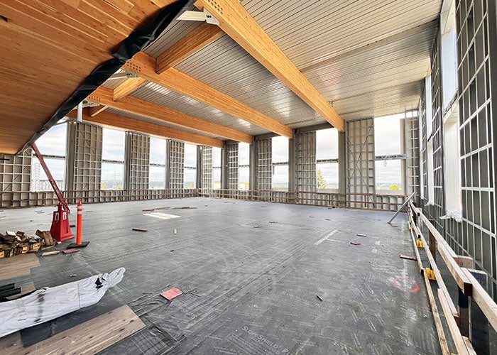 a large interior room under construction with part of the ceiling wood and large wood beams. Windows are in all three walls that are showing. The walls are framed but do not have wallboard.