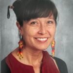 Benayshe Titus, a woman with a maroon colored shirt and dangly earrings smiles in front of a grey background.
