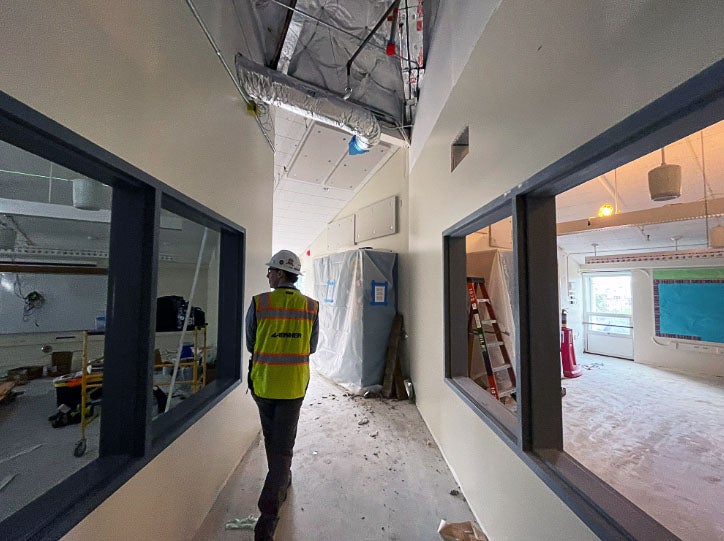 a person stands in a mostly complete hallway with windows on each side. the ceiling has ductwork showing and the floor is protected.
