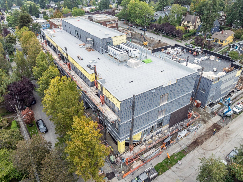 a large 3 story building under construction with gray material on the outside and trees next to it with green leaves