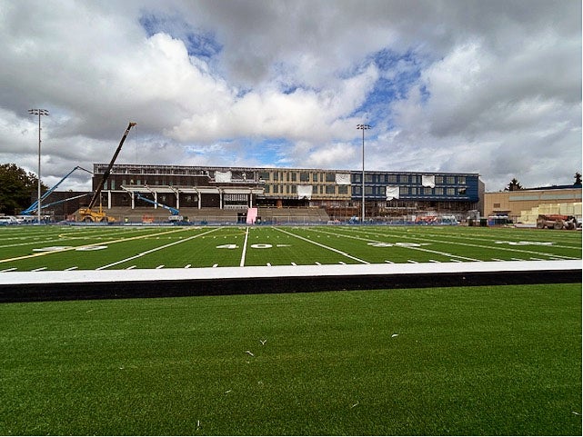 looking toward a building with construction equipment in front over a green turf field marked for football