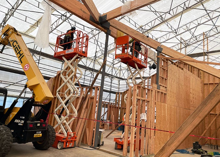 two scissor lifts with people and some ceiling beams above partially framed walls inside a white tent on scaffolding
