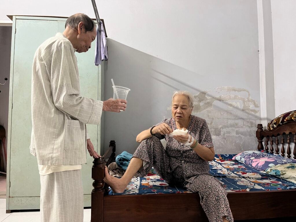 Two people sitting on a bed and standing while eating food in room with white walls