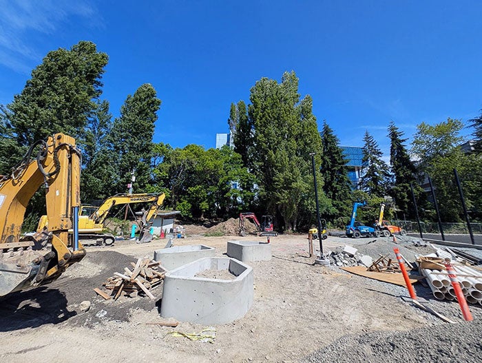 A dirt construction zone with multiple excavators and building materials scattered around. Large concrete forms and pipes are staged near the center, surrounded by tall trees and buildings in the background.